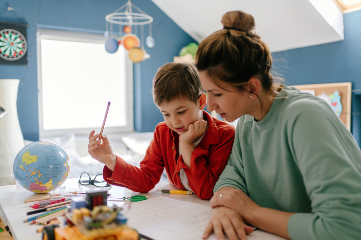 Abbildung zeigt Mutter, die mit ihrem Sohn im Kinderzimmer sitzt und Homeschooling-Aufgaben bearbeitet.