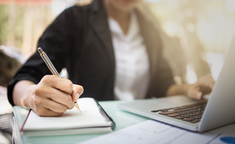 istock.com/iBrave Abbildung zeigt Lehrerin im Hintergrund, die im Vordergrund auf einem Laptop arbeitet und sich gleichzeitig auf einem Notizblock Bemerkungen notiert.